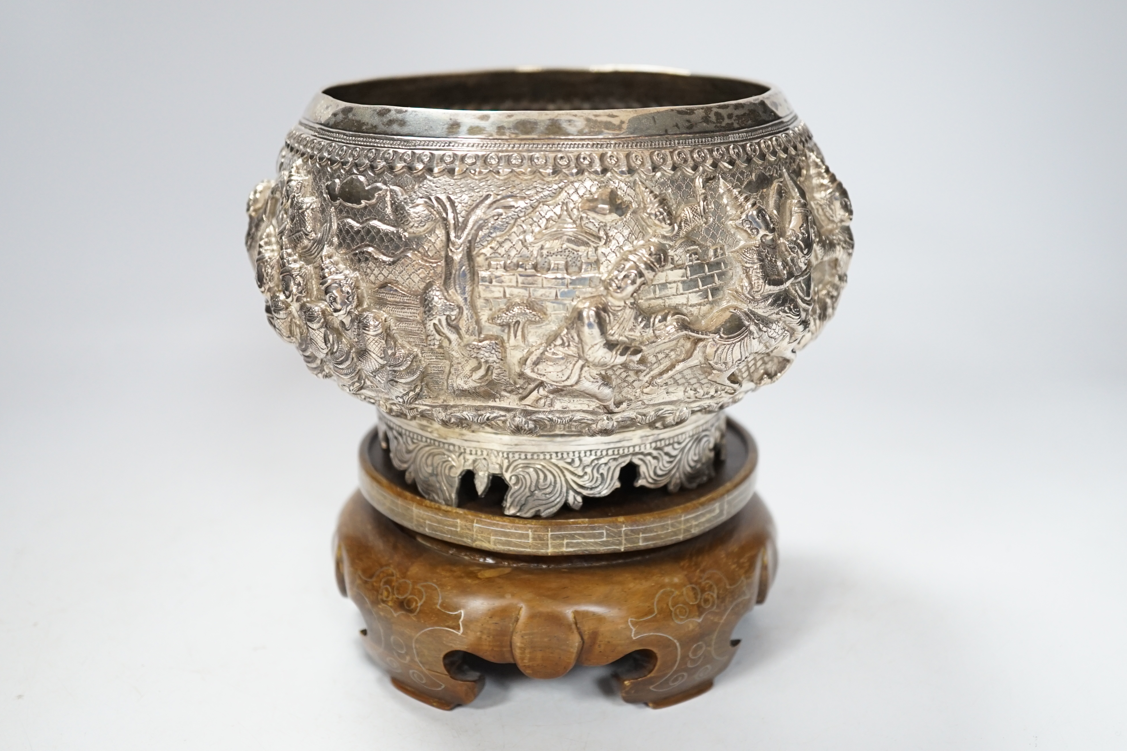 An Indian circular white metal bowl, decorated with a continuous band of figures in a landscape, on scrolled feet with hardwood stand, diameter 16cm